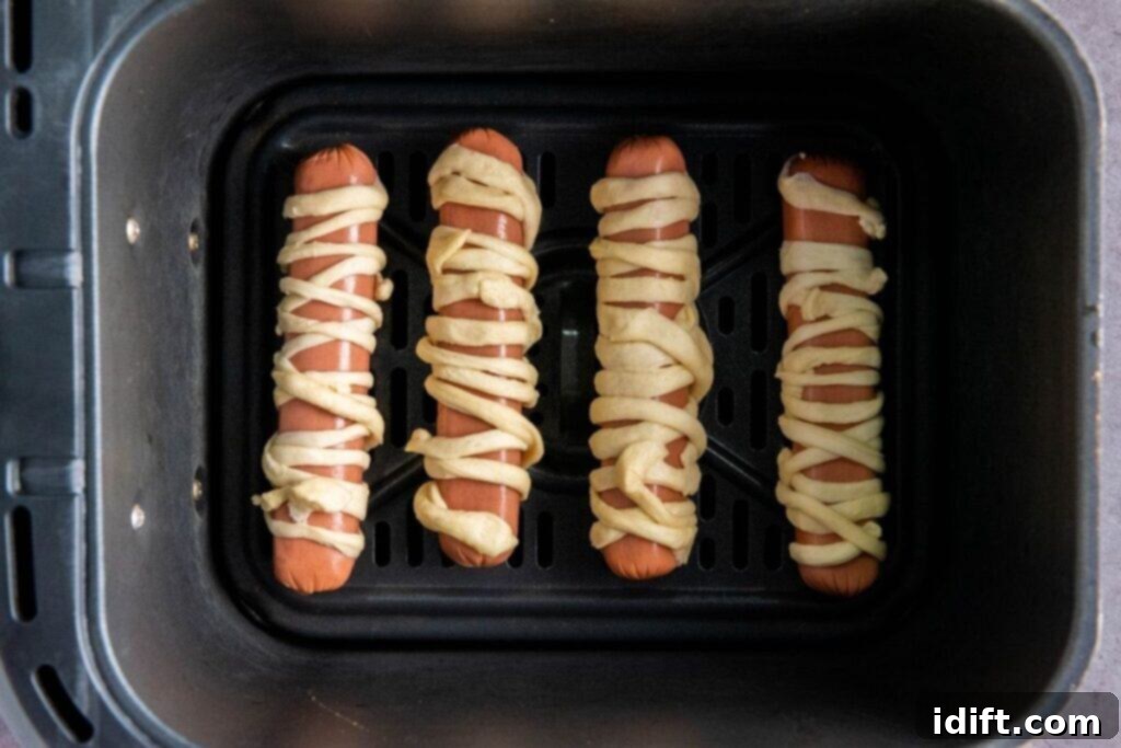 Four hot dogs, meticulously wrapped in crescent dough to resemble mummies, are neatly arranged in a single layer within an air fryer basket, ready for cooking. The spacing allows for optimal air circulation.