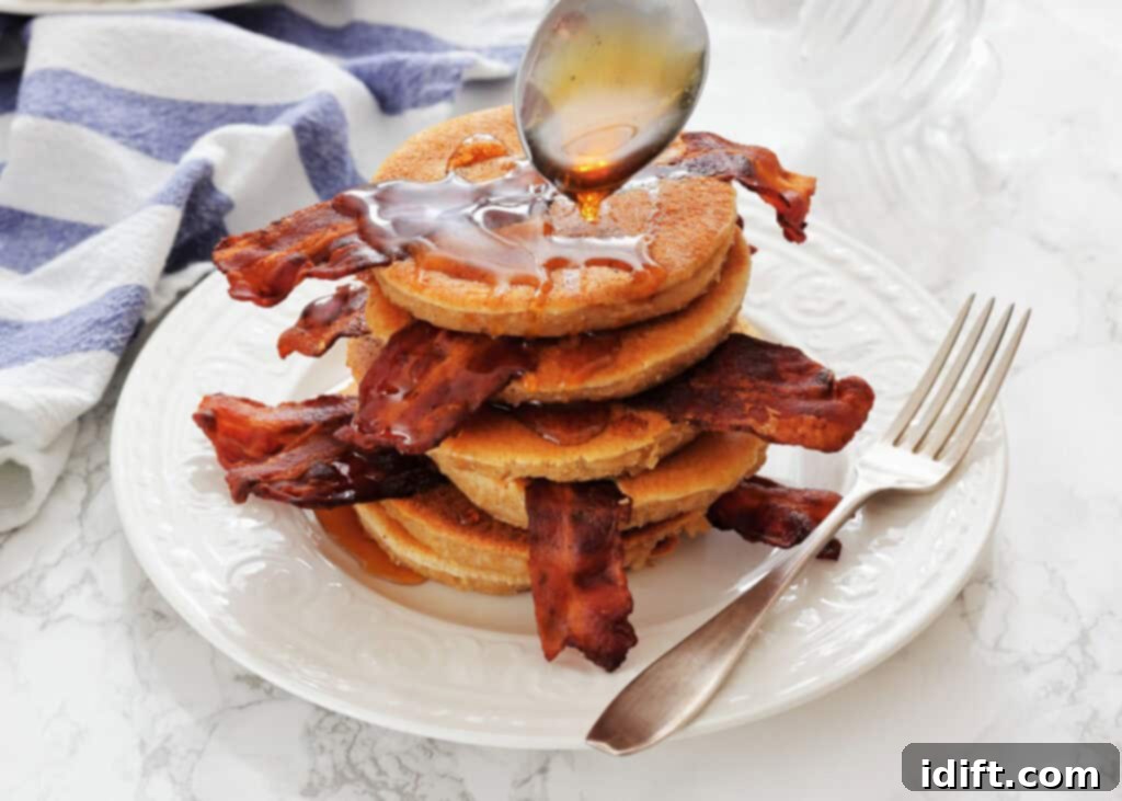 Stacked on Bacon Pancakes on a white plate, being drizzled with syrup from a spoon, with a fork beside the stack.