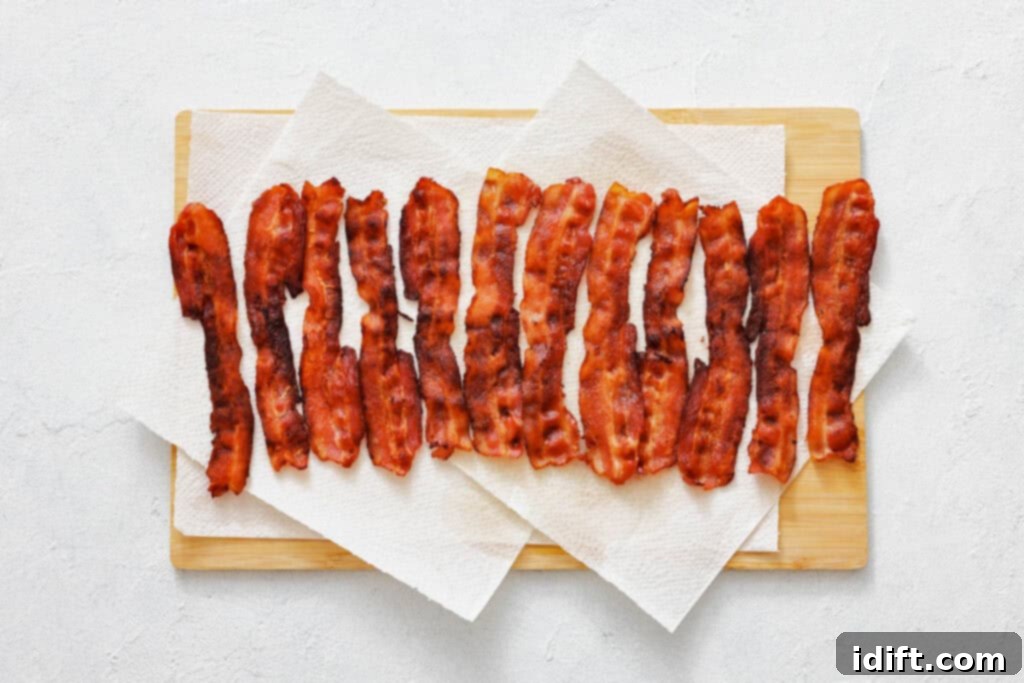 Crispy strips of cooked bacon are arranged in a row on paper towels placed on a wooden cutting board.