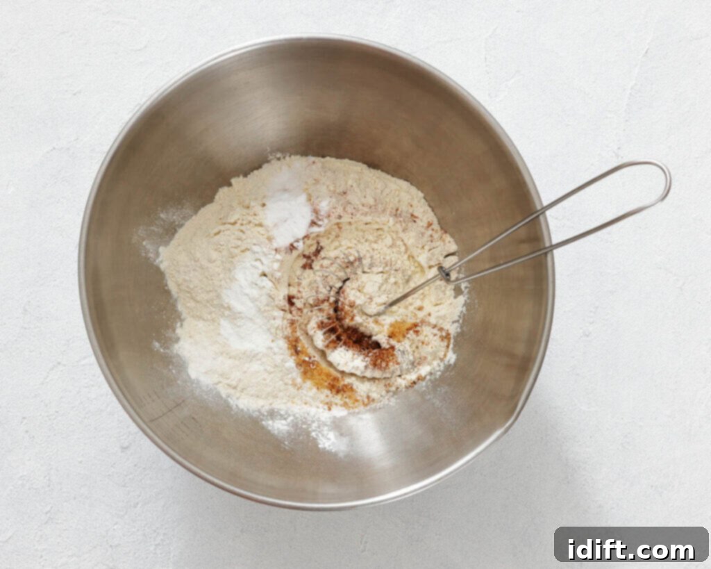 A metal mixing bowl contains flour, baking soda, baking powder, cinnamon, and other dry ingredients, with a metal whisk resting inside.