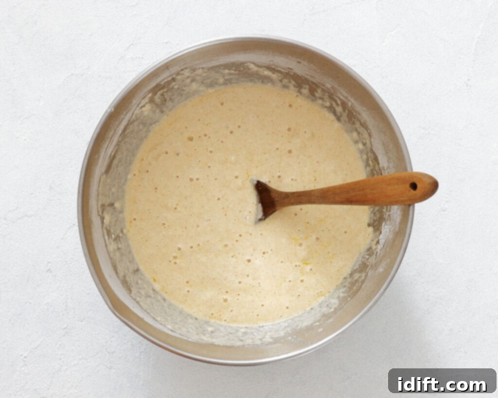 A stainless steel bowl filled with pancake batter sits on a white surface with a wooden spoon partially submerged in the mixture.