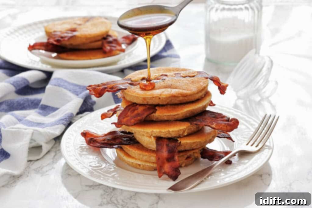 A stack of Bacon Pancakes on a white plate, with syrup being poured on top and a fork beside the plate. A jar of milk and another plate are in the background.