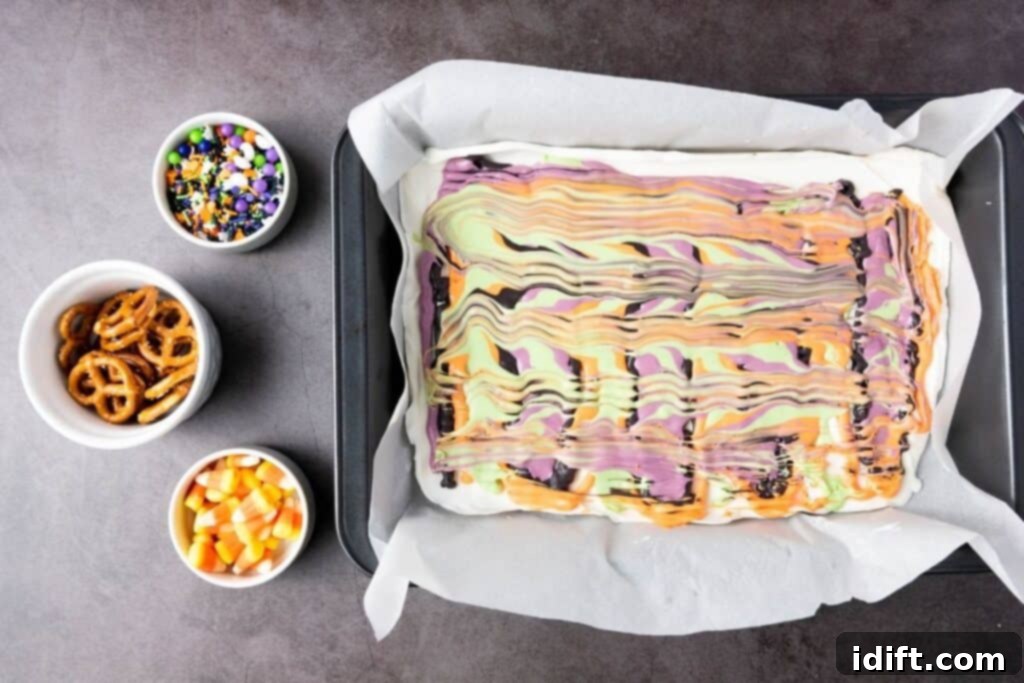 A baking tray with swirled Halloween-colored bark on parchment paper, next to small bowls of Halloween sprinkles, pretzels, and candy corn.