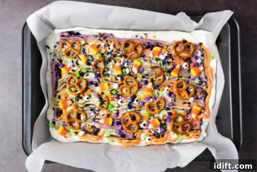 A tray of Halloween-themed dessert bark topped with pretzels, candy corn, colorful drizzles, and festive sprinkles on parchment paper.