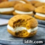 Fluffy Pumpkin Whoopie Pies with Cream Cheese Frosting 12 Pumpkin whoopie pie with cream filling on a metal plate, partially eaten to reveal its soft, center. More pies are blurred in the background.