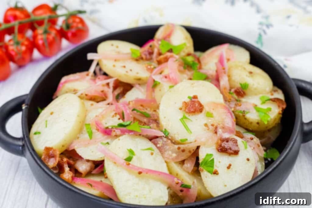 A black dish filled with sliced potatoes, red onions, bacon pieces, chopped parsley, and mustard seeds. Cherry tomatoes are in the background.