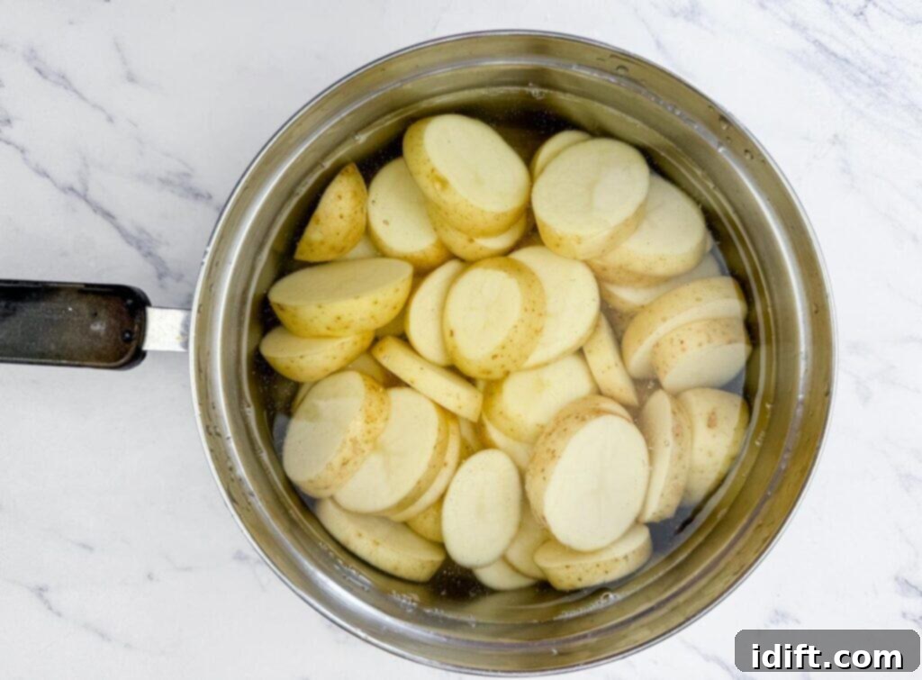 A stainless steel pot filled with sliced potatoes submerged in water, placed on a white marble surface.