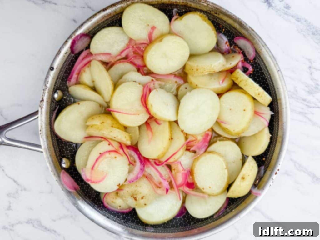 Sliced potatoes and red onions cooking in a stainless steel skillet on a white marble surface.