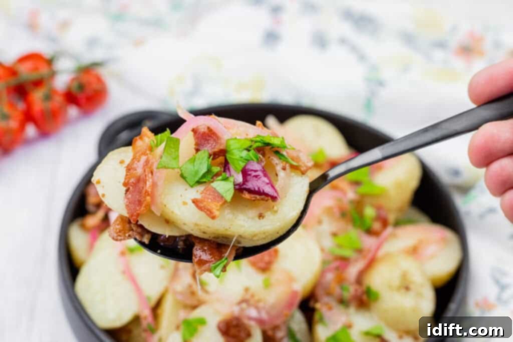 A spoonful of sliced potato salad with bacon, onions, and chopped parsley, held above a bowl of the same dish.
