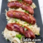 Four grilled sausages are served on a bed of sauerkraut and garnished with chopped herbs on a black rectangular plate.