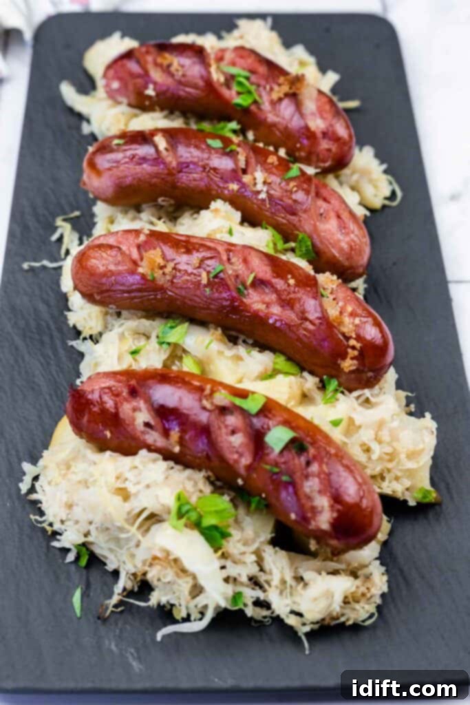 Four grilled sausages are served on a bed of sauerkraut and garnished with chopped herbs on a black rectangular plate.