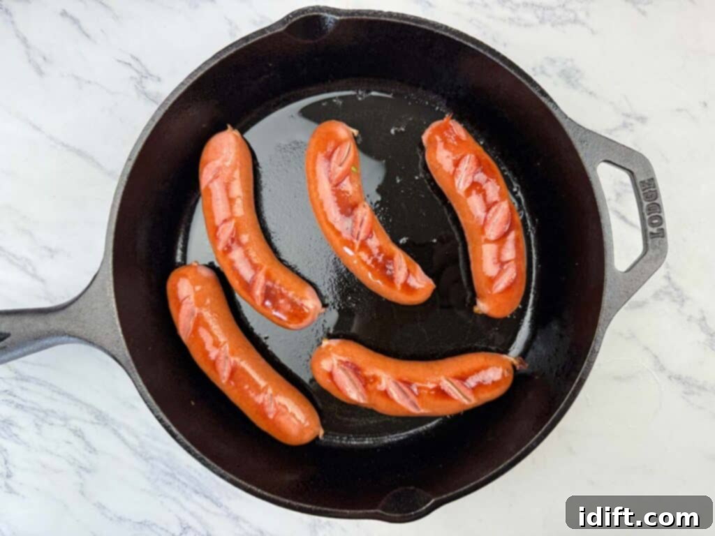 Five scored hot dogs are cooking in a black cast iron skillet on a white marble surface.