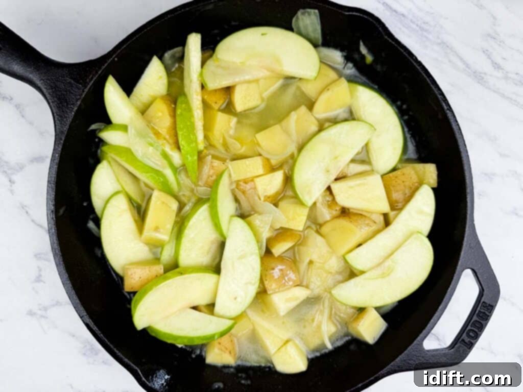 A cast iron skillet containing sliced green apples and chopped potatoes cooking in a light sauce on a marble countertop.