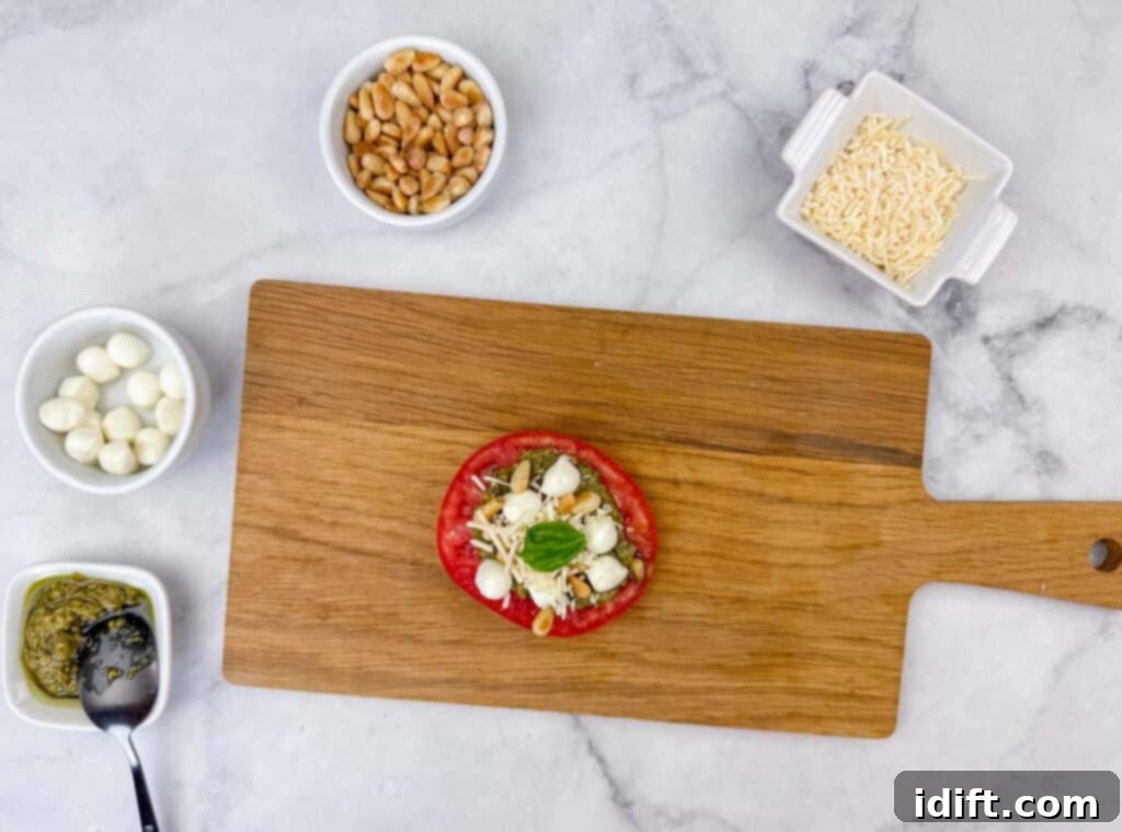 A rustic wooden cutting board displays a fresh tomato slice beautifully topped with creamy cheese, tender mozzarella balls, toasted pine nuts, and fragrant basil. Surrounding it are small, elegant bowls holding additional pine nuts, grated cheese, more mozzarella balls, and a rich basil pesto, all ready for a culinary creation.