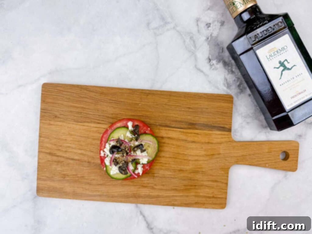 A rustic wooden cutting board showcases a vibrant tomato slice expertly topped with crisp cucumber, creamy feta, savory olives, and aromatic herbs. A bottle of elegant olive oil is positioned nearby on a sleek marble countertop, enhancing the culinary presentation.