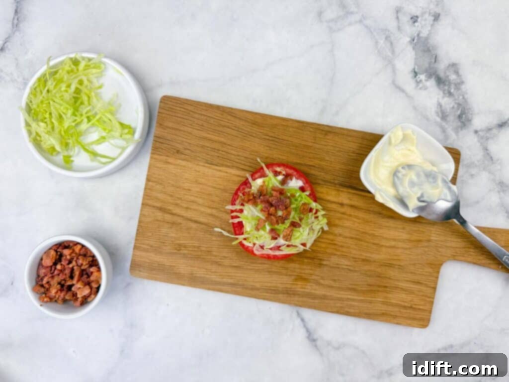 A rustic wooden board presents a vibrant tomato slice meticulously topped with shredded lettuce and crispy bacon bits. Adjacent to it, small bowls filled with fresh lettuce, savory bacon bits, and a delicate dish of creamy mayonnaise with a spoon are artfully arranged on a polished marble surface, creating an inviting culinary scene.