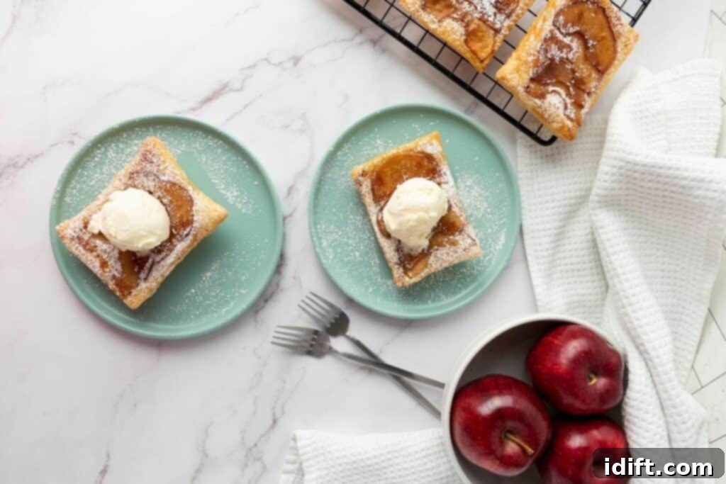 Two plates with apple tarts topped with ice cream, a cooling rack with more tarts, and a bowl of red apples on a white marble surface.