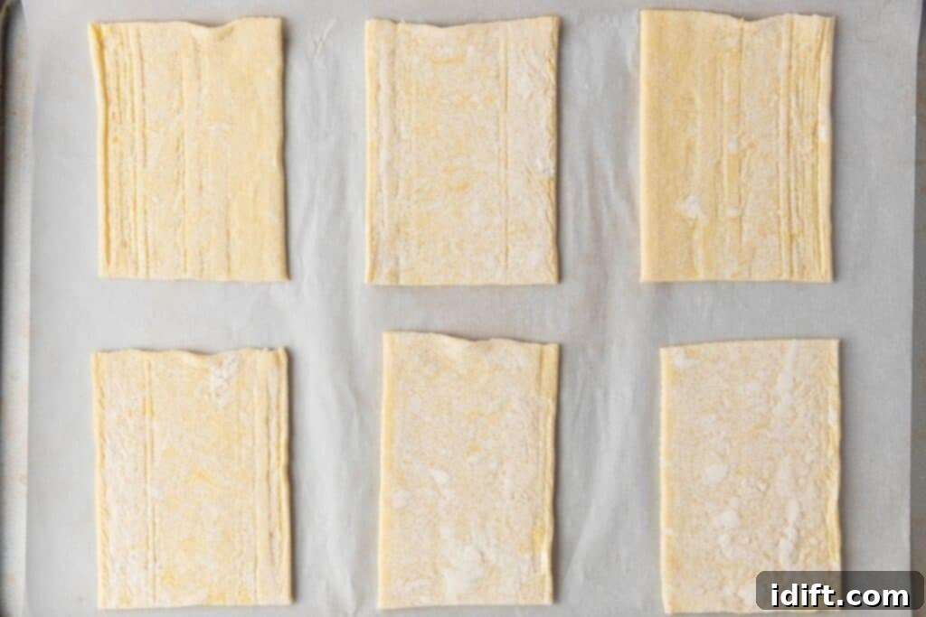 Six rectangular pieces of raw puff pastry dough are arranged in two rows on a parchment-lined baking sheet.