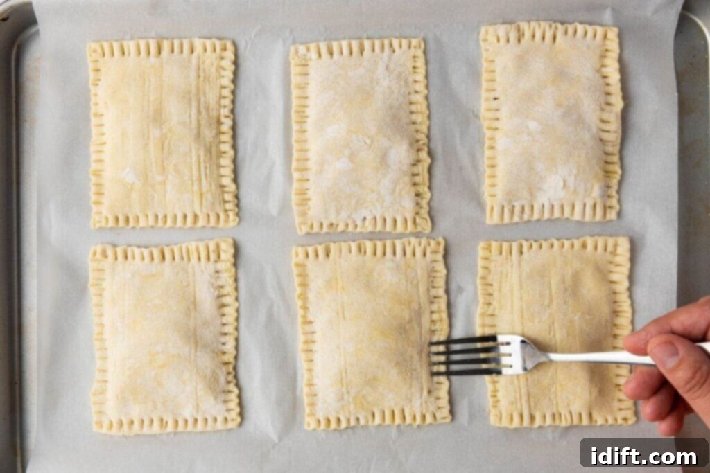 Six rectangular pastry pockets on parchment paper, with a hand using a fork to crimp the edges of one of them.