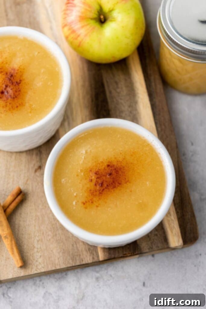 Chef Jenn's Effortless Instant Pot Applesauce 2 Two bowls of applesauce topped with cinnamon sit on a wooden board, next to an apple, a jar of applesauce, and two cinnamon sticks.