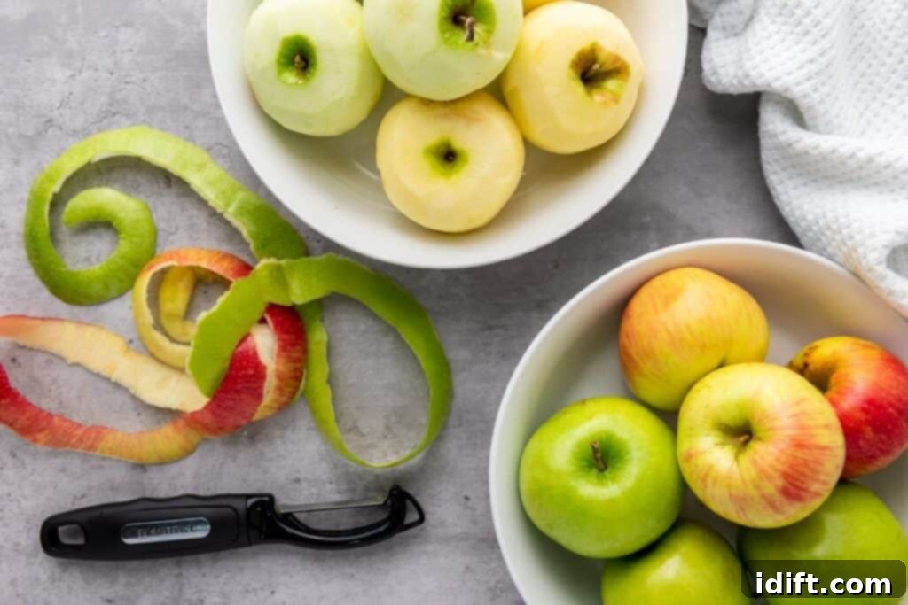 Chef Jenn's Effortless Instant Pot Applesauce 5 Two bowls of apples, one with peeled apples and one with unpeeled apples, sit on a gray surface beside apple peels, a peeler, and a white textured cloth.