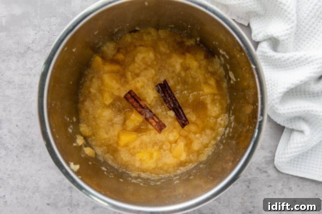 Chef Jenn's Effortless Instant Pot Applesauce 8 A stainless steel pot containing chunky applesauce with two cinnamon sticks, placed on a light gray surface next to a white textured cloth.