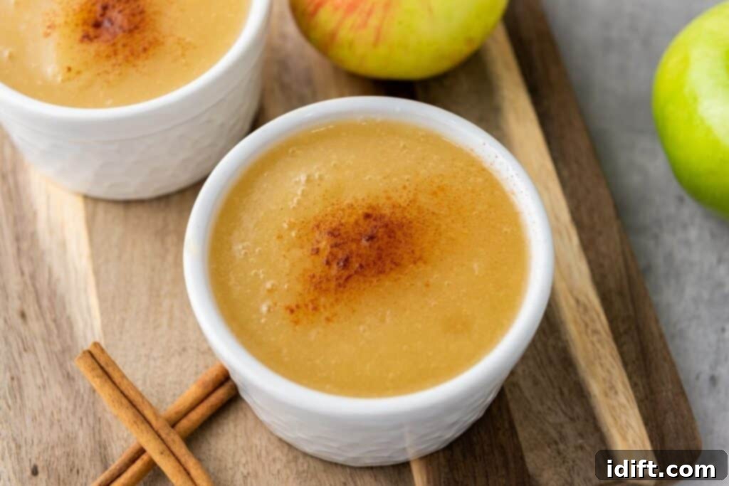 Two white bowls of applesauce topped with ground cinnamon, placed on a wooden board with fresh green apples and cinnamon sticks nearby.