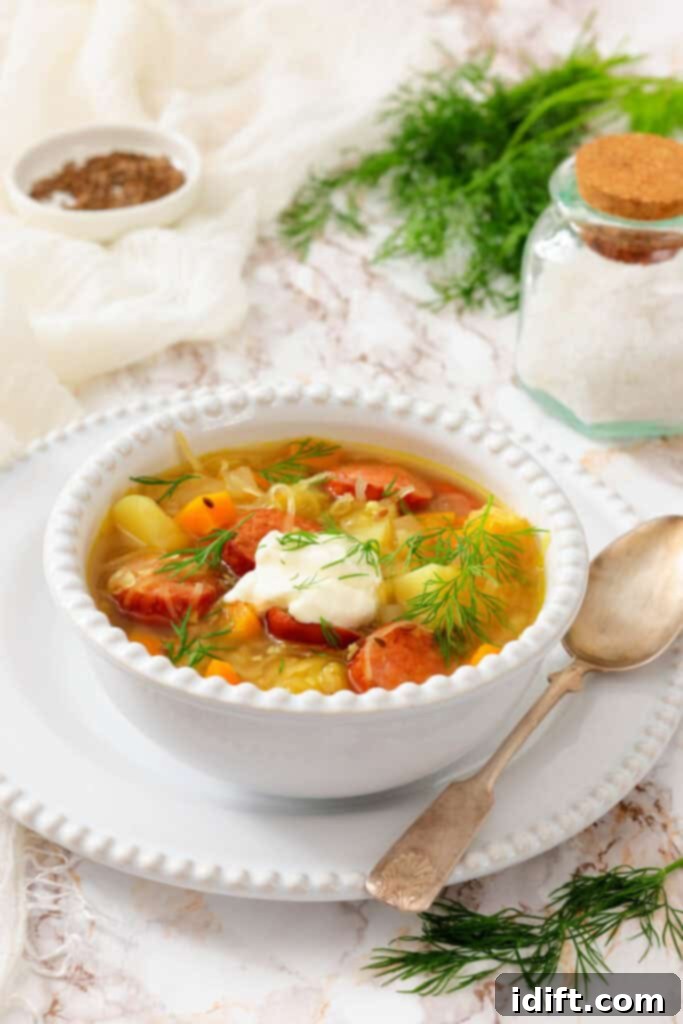 A bowl of vegetable and sausage soup topped with sour cream and fresh dill, accompanied by a spoon and surrounded by salt, pepper, and herbs on a white table.