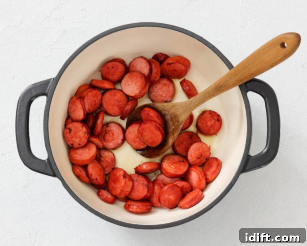 Sliced sausage pieces are being cooked in a white pot with a wooden spoon on a light surface.