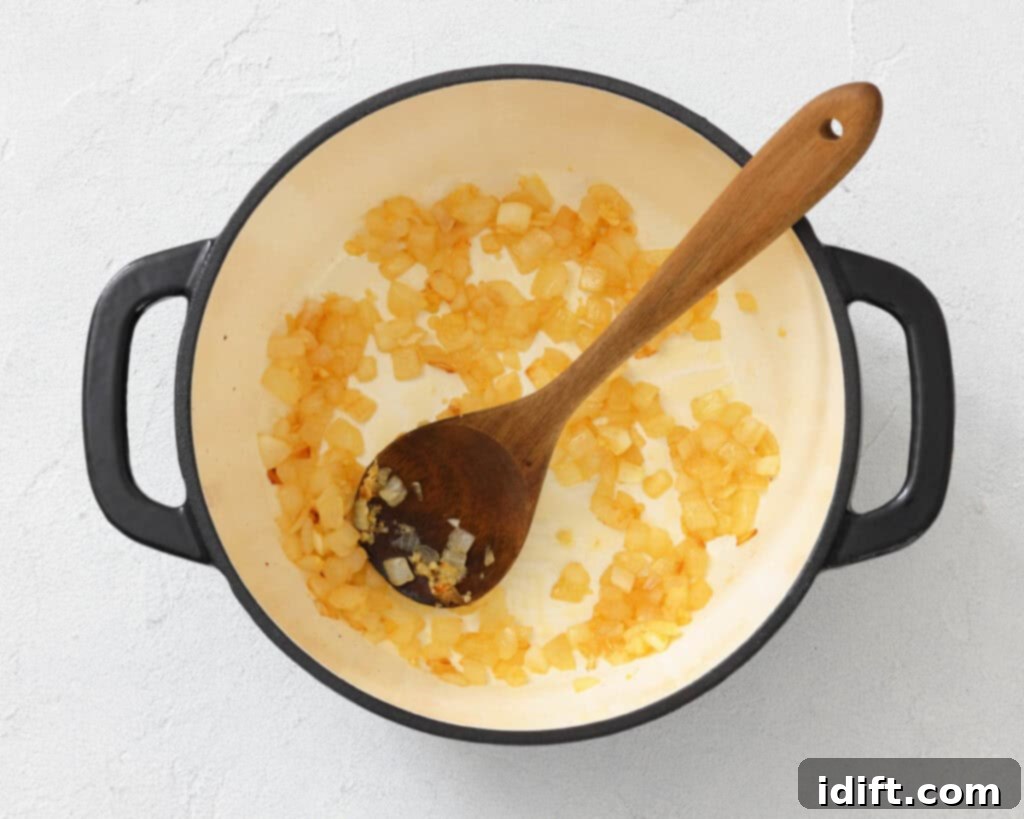 Diced onions sautéing in a white pot with a wooden spoon, placed on a light-colored surface.