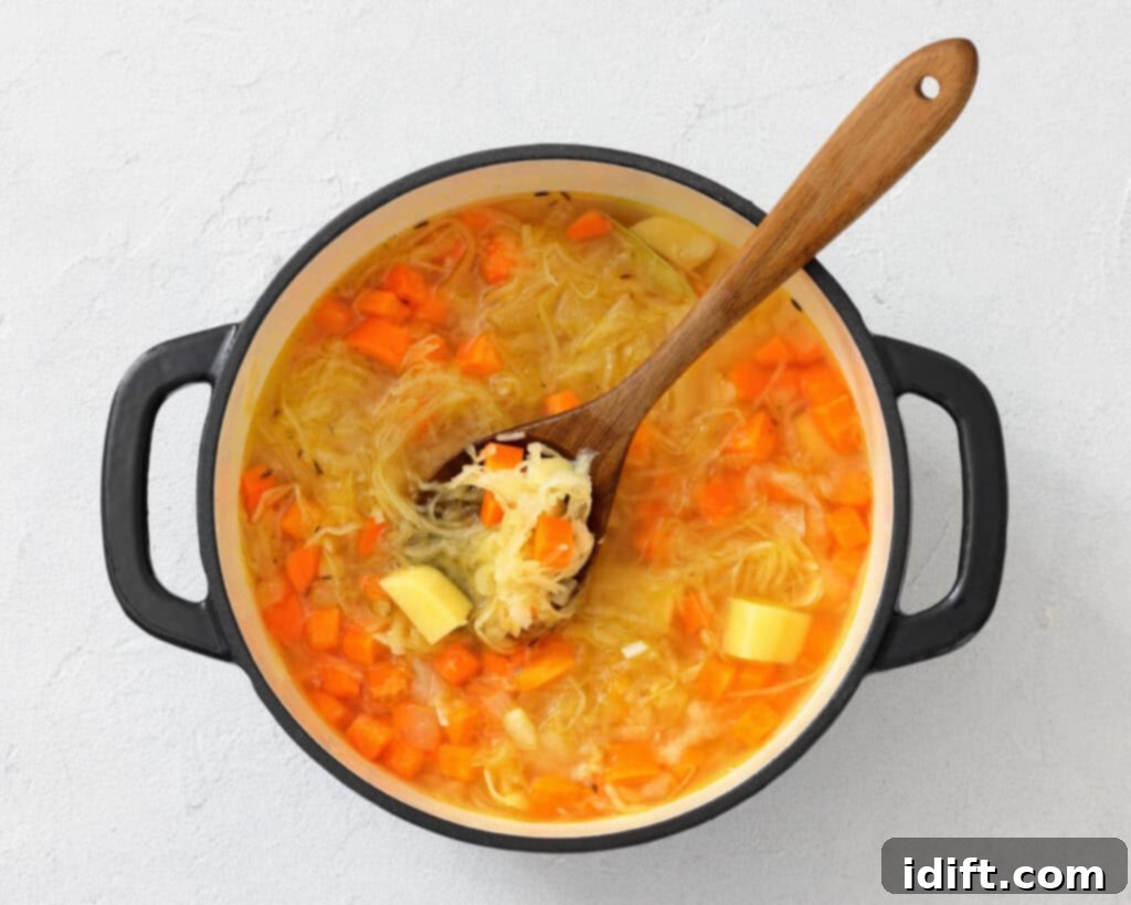 A pot of vegetable soup with carrots, potatoes, and shredded cabbage is being stirred with a wooden spoon.