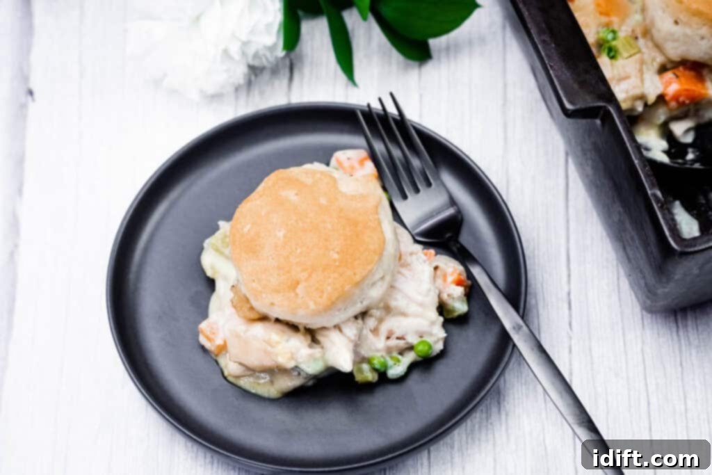 A black plate with a serving of chicken pot pie topped with a biscuit, a fork, and a casserole dish with more pot pie partially visible.