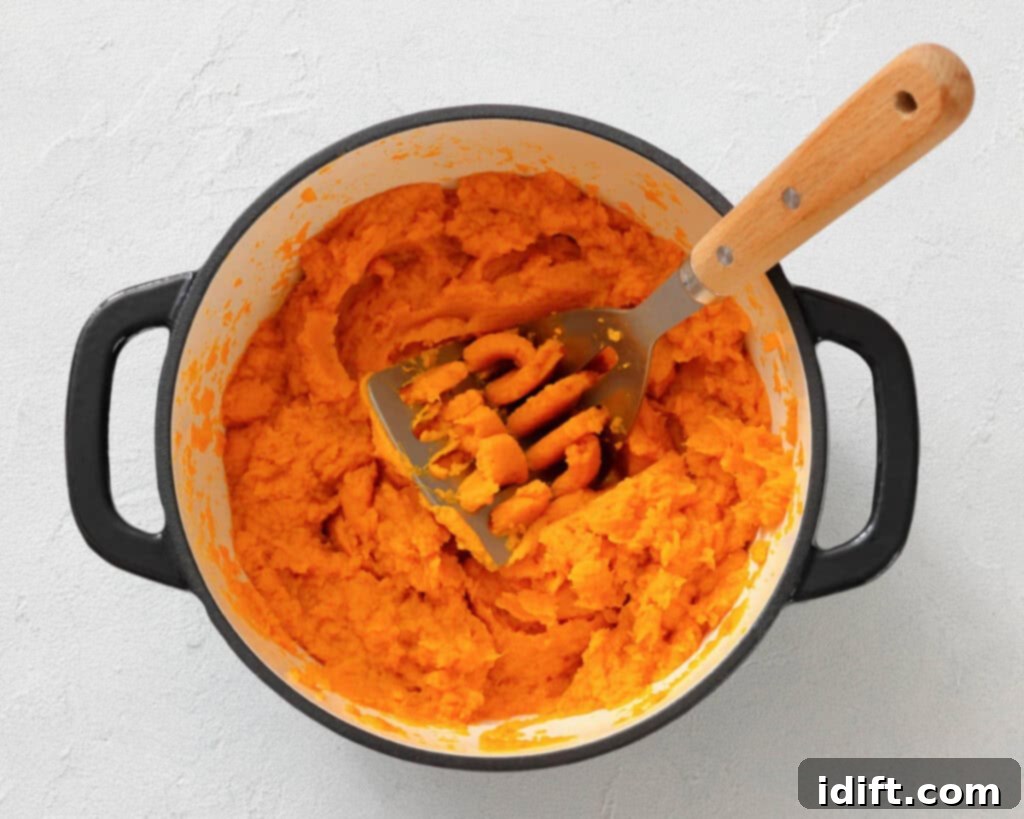 Decadent Sweet Potato Casserole with Crunchy Pecan Streusel 5 A pot of mashed sweet potatoes with a metal masher resting inside on a light-colored surface.